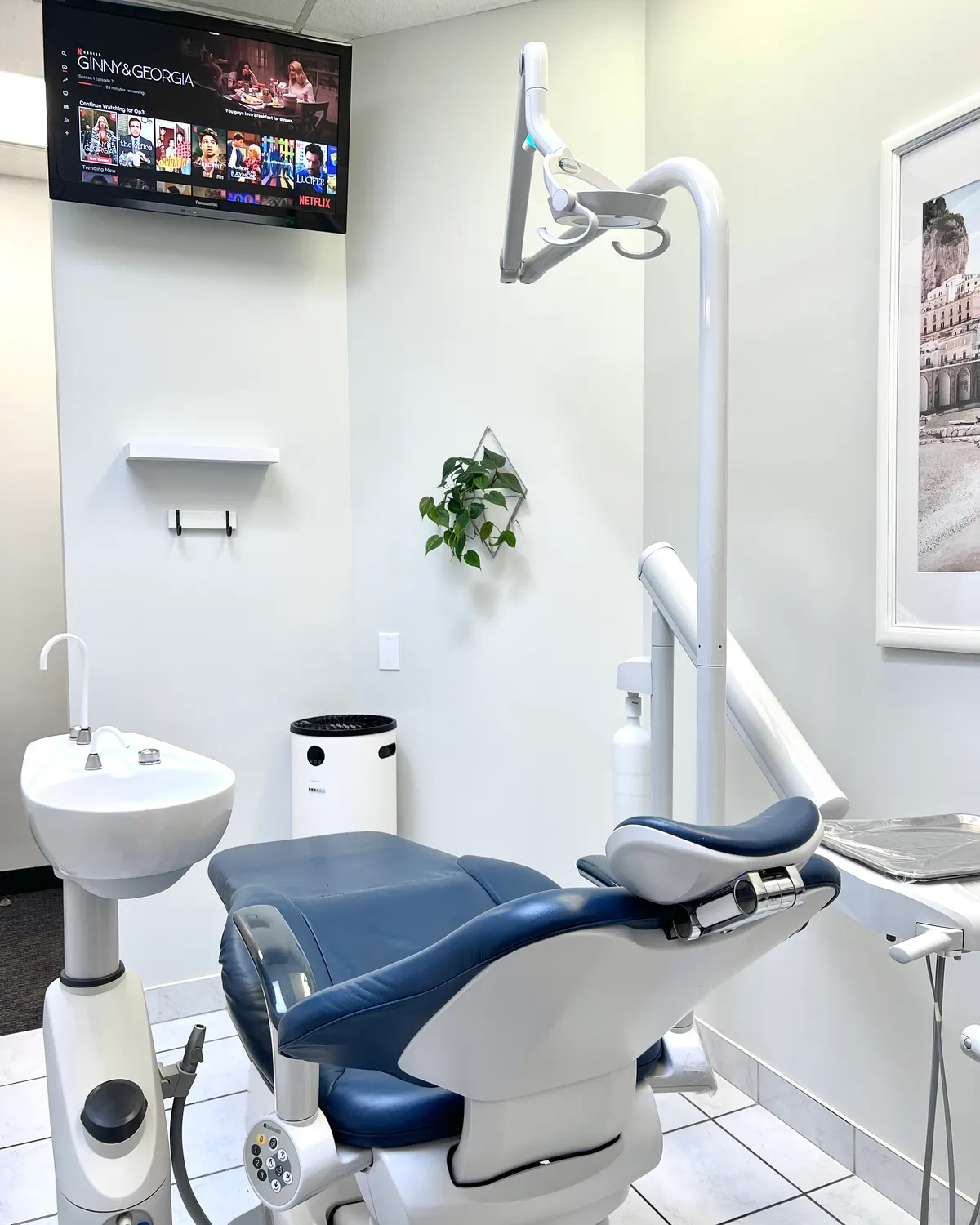 Headon treatment room with a navy dental chair, sink, overhead light and a wall-mounted TV